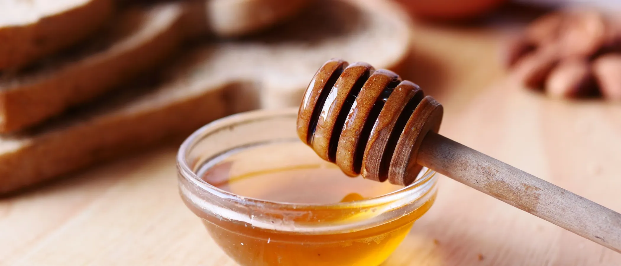 Close-up of honey dipper in a bowl with honey on wooden table beside sliced bread.